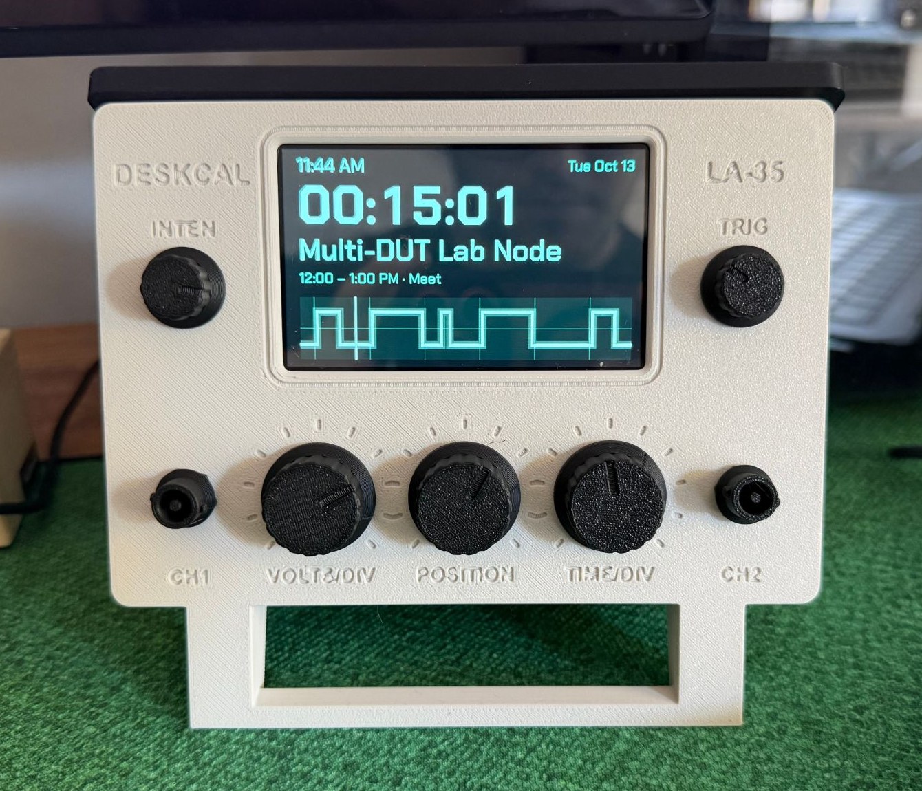 A cream 3D-printed stand shaped like a small oscilloscope, with black knobs and a handle, its screen counting down fifteen minutes to the next meeting above a waveform of the day